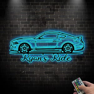 May include: A neon blue car outline with the text "Ryan's Ride" against a dark brick wall. The car is a coupe with detailed wheels and a spoiler. A remote control is held in the bottom right corner.