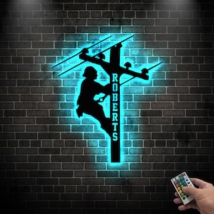 May include: A black metal silhouette of a lineman climbing a utility pole with the name "ROBERTS" on the pole. The silhouette is illuminated with a bright blue light.