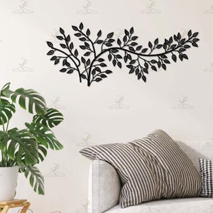 May include: Black metal wall art depicting a stylized tree branch with numerous leaves. The artwork is mounted on a white wall above a sofa with striped and checkered pillows. A potted plant with large green leaves is in the foreground.