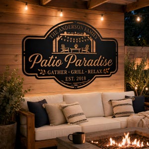 Puede incluir: Letrero de metal negro con las palabras "Patio Paradise" y "The Anderson Family" en una fuente decorativa. El letrero también incluye las palabras "GATHER. GRILL. RELAX." y "EST. 2018". El letrero está montado en una pared de madera.