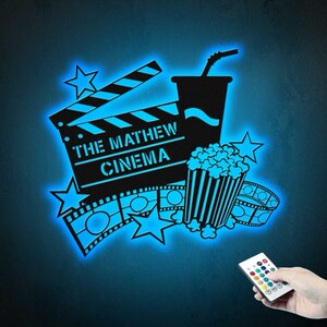 May include: A blue glow-in-the-dark wall decal featuring a movie clapperboard, popcorn bucket, and film reel. The clapperboard reads "The Mathew Cinema".
