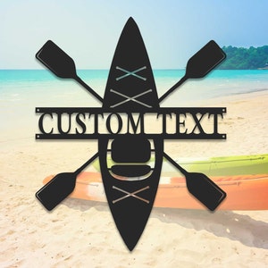 May include: Black metal silhouette of a kayak with two crossed paddles and the text "CUSTOM TEXT" on a wooden sign. The silhouette is in front of a beach scene with a kayak in the sand.