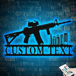 May include: Black metal silhouette of a rifle with a scope and bullets, mounted on a shelf with the words "CUSTOM TEXT" in blue neon lights. A remote control with colored buttons is in the lower right corner.