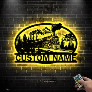 Puede incluir: Una silueta de metal de un tren que viaja a través de un paisaje montañoso con árboles a cada lado. La silueta está iluminada con luces amarillas y las palabras "CUSTOM NAME" se muestran debajo del tren.