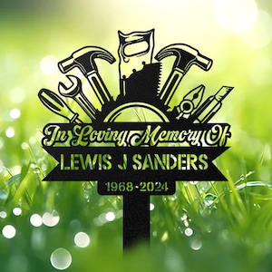 May include: Black metal garden memorial featuring tools such as a hammer, saw, wrench, and screwdriver. The memorial reads "In Loving Memory Of LEWIS J SANDERS 1968-2024".