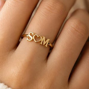 Op de afbeelding: Een goudkleurige ring met de letters S en M gescheiden door een hartvorm. De tekst aan de rechterkant luidt WORN WITH Love, SUBTLE. PERSONAL. TIMELESS., DAILY WEAR, MINIMAL & ELEGANT, MEANINGFUL GIFT, MADE JUST FOR YOU, Carry your story with you, always.
