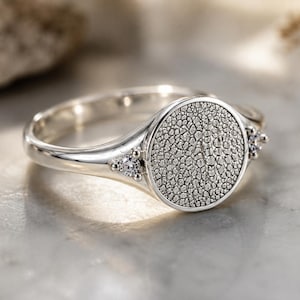 May include: A silver ring featuring a circular face with a textured, pebbled surface. The band is polished silver, and two small, clear, round-cut stones are set on either side of the central circular element, adding a subtle sparkle to the design.