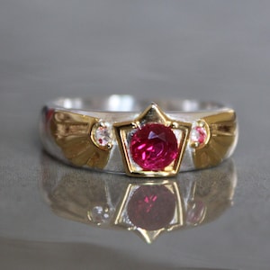 May include: A silver ring with a gold crown design featuring a red ruby gemstone and two smaller white gemstones. The ring has a wing-like design on either side of the crown.