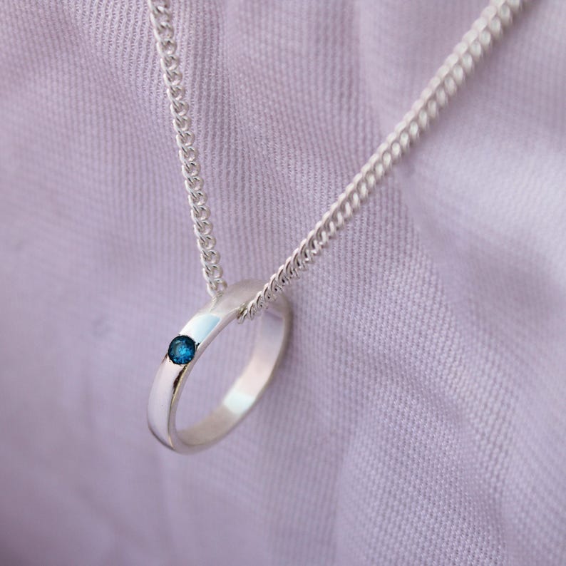 May include: A silver pendant necklace featuring a ring design with a small, round, blue gemstone. The ring is attached to a delicate silver chain. The background is a soft, light purple fabric.
