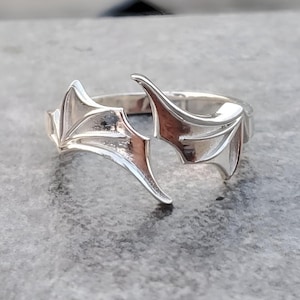 Może przedstawiać: Srebrny pierścionek o unikalnym wzorze z dwoma stylizowanymi skrzydłami. Skrzydła są otwarte i zakrzywione, tworząc delikatny i misterny wygląd.