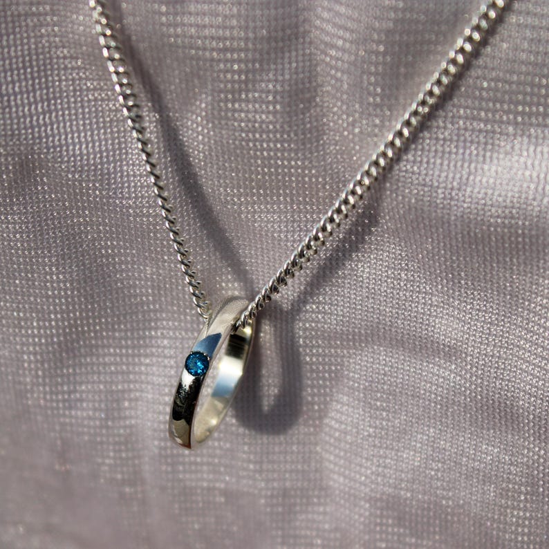 May include: A silver ring pendant necklace with a small, round, blue gemstone set into the ring. The ring hangs from a delicate silver chain. The background is a soft, textured, light gray fabric.