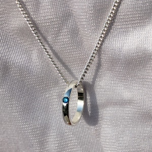 May include: A silver pendant necklace featuring a ring design with a small, round, blue gemstone. The pendant hangs from a delicate silver chain against a soft, textured background.