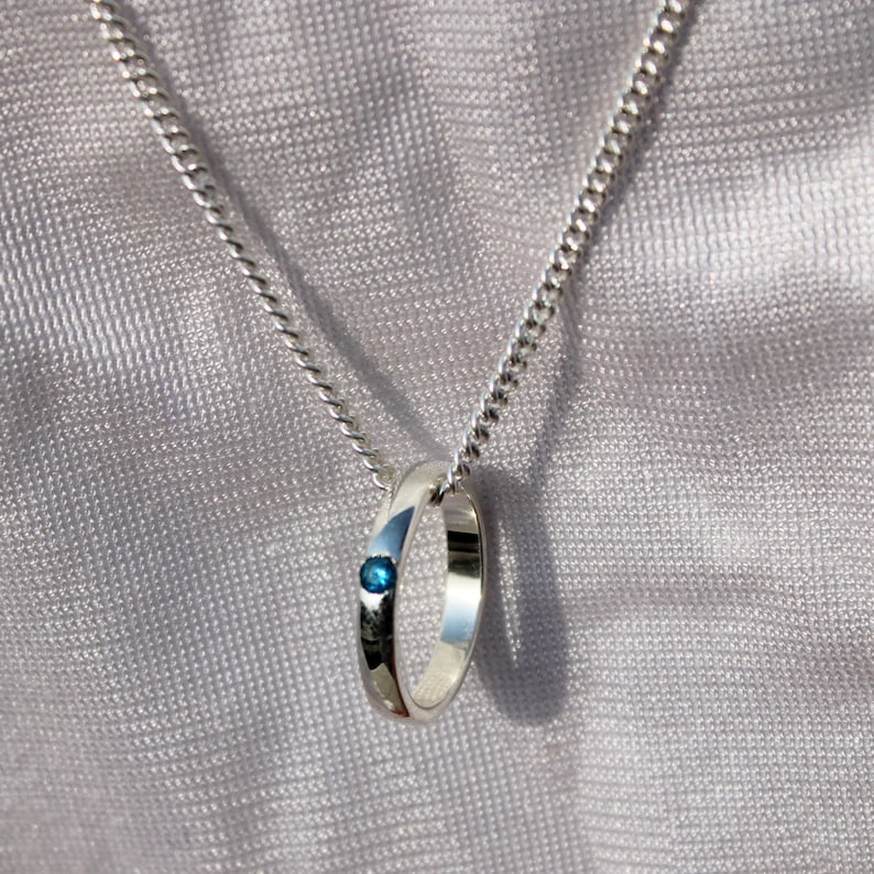 May include: A silver pendant necklace featuring a ring design with a small, round, blue gemstone. The pendant hangs from a delicate silver chain against a soft, textured background.