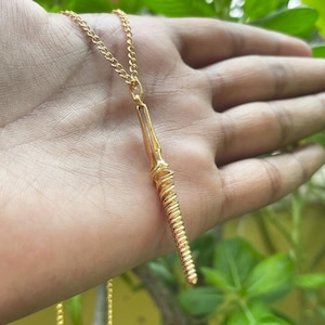 May include: A gold-colored pendant necklace. The pendant has a long, thin, vertical design with a spiral wire wrapping around the lower portion. The necklace chain is a delicate gold color. The background is a hand and green foliage.