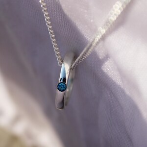 May include: A silver pendant necklace featuring a circular design with a small, round, blue gemstone. The pendant hangs from a delicate silver chain. The jewelry is displayed against a soft, white background.