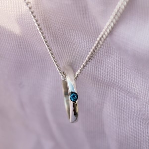 May include: A silver pendant necklace featuring a ring-shaped charm with a small, round, blue gemstone. The necklace has a delicate chain and is displayed against a soft, light background.