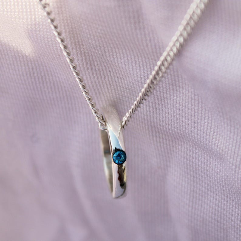 May include: A silver pendant necklace featuring a ring-shaped charm with a small, round, blue gemstone. The necklace has a delicate chain and is displayed against a soft, light background.