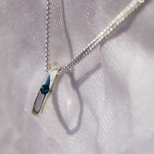 May include: A silver pendant necklace with a small, round, blue gemstone set in a rectangular silver pendant. The pendant hangs from a delicate silver chain. The background is a soft, white fabric.