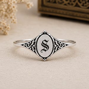May include: A silver ring featuring an oval-shaped centre plate engraved with the letter S in a gothic font. The band and the setting are decorated with intricate, dark-oxidised scrollwork patterns that extend from the central oval element.