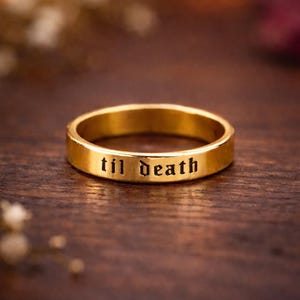 Könnte beinhalten: Ein goldfarbener Ring mit der Inschrift "til death" in schwarzer, gotischer Schrift. Der Ring ist vor einem verschwommenen, dunklen Holzgrund platziert, wodurch die Inschrift und das Design des Rings hervorgehoben werden.