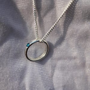 May include: A silver ring pendant necklace with a small, round, blue gemstone. The pendant is attached to a delicate silver chain. The necklace is displayed against a soft, textured, gray background.