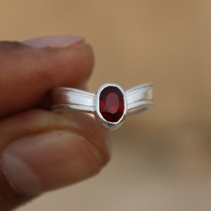 Może przedstawiać: Srebrny pierścionek z czerwonym, owalnym kamieniem szlachetnym osadzonym w środku. Pierścionek ma prosty design z dwoma zakrzywionymi liniami po obu stronach kamienia.