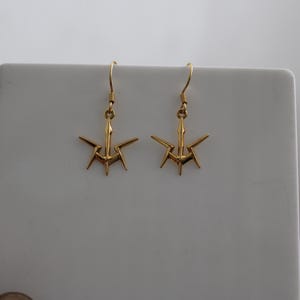 Puede incluir: Un par de pendientes dorados. Cada pendiente presenta un diseño estilizado en forma de estrella con múltiples puntas que se extienden hacia afuera desde un elemento central. Los pendientes tienen un cierre de gancho para orejas perforadas.