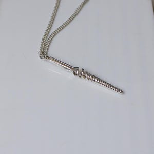 May include: A silver-colored pendant necklace featuring a stylized, screw-shaped design. The pendant has a detailed, spiral-like texture and is suspended from a delicate chain. The necklace is displayed on a white surface.