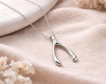 May include: A silver-colored wishbone pendant necklace. The pendant is smooth and polished, suspended from a delicate chain. The necklace is displayed on a soft, peach-colored fabric, with blurred floral accents in the background.