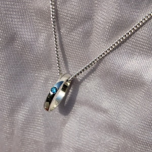 May include: A silver pendant necklace featuring a ring-shaped charm with a small, round, blue gemstone. The pendant hangs from a delicate silver chain. The background is a soft, textured, gray fabric.
