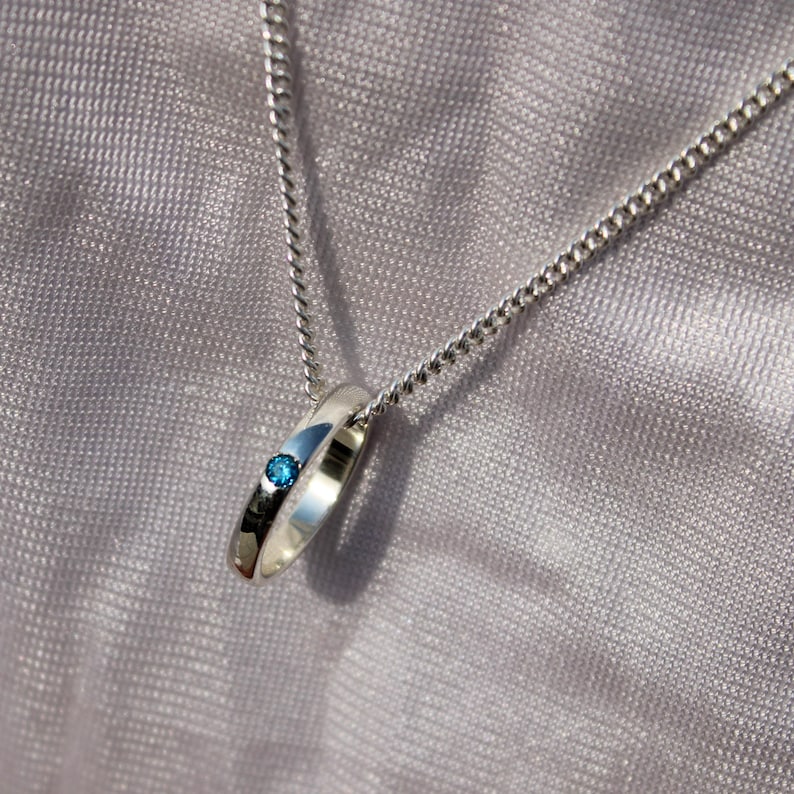 May include: A silver pendant necklace featuring a ring-shaped charm with a small, round, blue gemstone. The pendant hangs from a delicate silver chain. The background is a soft, textured, gray fabric.