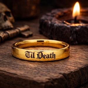 May include: A gold-colored ring with the inscription "Til Death" in a gothic font. The ring is centered on a wooden surface, with a skull, keys, and a lit candle in the background. The ring is a simple band.