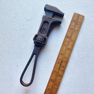 May include: An antique adjustable wrench and a wooden ruler. The wrench is made of dark metal and shows signs of wear. The ruler is wooden with inch markings. The items are placed on a white surface.