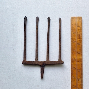 May include: A rusty, four-tined garden fork with a single central tine. The fork is made of iron and is next to a wooden ruler. The tines are approximately 6 inches long.