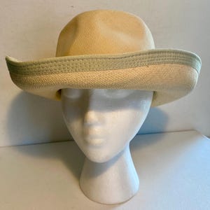 May include: A straw hat with a light beige crown and a wide brim. The brim is trimmed with a light green band. The hat is displayed on a white mannequin head. This hat is perfect for sun protection and fashion.