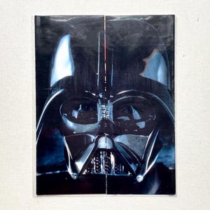 May include: A close-up image of Darth Vader's helmet, a prominent character from the Star Wars franchise. The helmet is predominantly black with reflective surfaces, and the image is set against a dark background. The image is a close-up of the character's face.