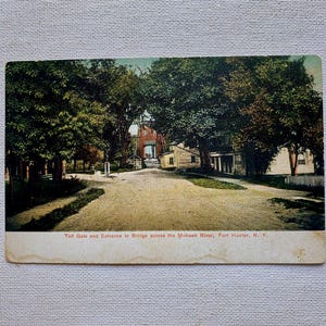 May include: Vintage postcard depicting the Toll Gate and Entrance to Bridge across the Mohawk River in Fort Hunter, N.Y. The image shows a dirt road leading to a brick building, framed by lush green trees. The text at the bottom reads "Toll Gate and Entrance to Bridge across the Mohawk River, Fort Hunter, N. Y."