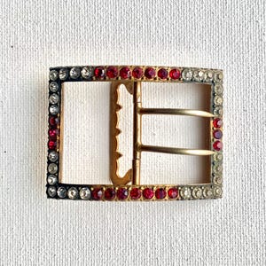May include: Rectangular vintage buckle with a gold-tone frame. The frame is encrusted with alternating red and clear rhinestones. The buckle has a gold-tone prong and a white backing.