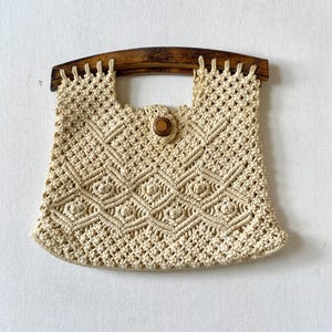 May include: A crocheted beige bag with a wooden handle and a wooden button closure. The bag has a diamond pattern.
