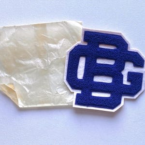 May include: A blue and white embroidered patch with the letters "BG" in a collegiate style. The patch is on a white background, next to a piece of translucent paper.