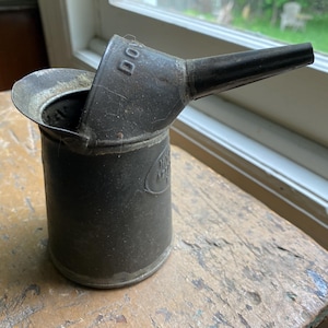 May include: An antique metal oil can with a spout and handle. The can is dark gray with visible wear and tear, suggesting age. The word "DO" is visible on the top. The can is sitting on a wooden surface.