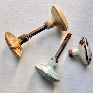 May include: Three vintage door knobs with weathered finishes. One knob is tan with dark rust, another is light blue with chipped paint, and the third is white with dark brown edges. Two knobs feature attached metal square spindles.