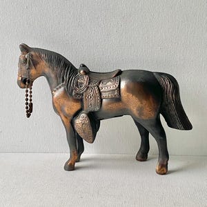 May include: A bronze-colored horse figurine with a saddle and bridle. The horse has a detailed mane and tail, and a beaded chain hangs from its mouth. The saddle is intricately designed, adding to the figurine's decorative appeal.