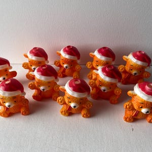 May include: Ten orange and red Christmas tree ornaments shaped like teddy bears wearing Santa hats.