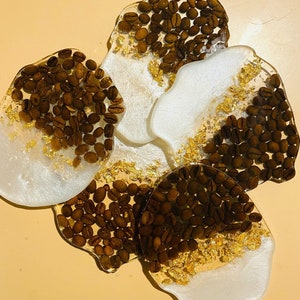 May include: Four white coasters with gold accents and brown coffee beans. The coasters are round and have a glossy finish.