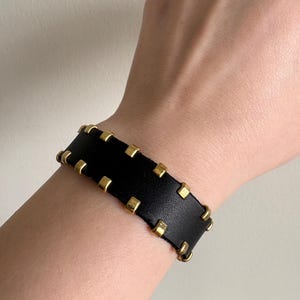 Puede incluir: Pulsera de cuero negro con tachuelas cuadradas doradas. La pulsera ancha es un accesorio atrevido. Las tachuelas doradas están espaciadas uniformemente alrededor de los bordes de la pulsera, añadiendo un toque de elegancia al diseño.