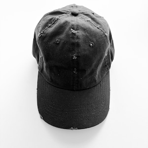 Może przedstawiać: Czarna, zniszczona czapka bejsbolówka ze lekko zakrzywionym daszkiem.