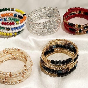 Puede incluir: Una variedad de pulseras de espiral con cuentas en varios colores y diseños. Una pulsera presenta un arcoíris de cuentas, mientras que otras muestran cuentas plateadas, rojas, perlas, doradas y negras. Estas pulseras son un accesorio elegante.