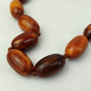 May include: A necklace featuring smooth, oval-shaped beads in varying shades of amber, brown, and dark mahogany. The beads are strung together with a thin, brown cord, with visible knots between each individual bead along the strand.