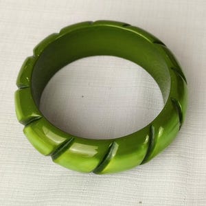 Può includere: Un braccialetto a cerchio verde lucido. Il braccialetto ha una forma arrotondata con una serie di rientranze rettangolari scolpite attorno all'esterno. Il colore è un verde primaverile vibrante e la superficie riflette la luce.
